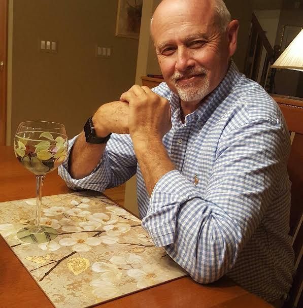 A man in a blue checkered shirt smiles at the camera while sitting at a table with a drink.