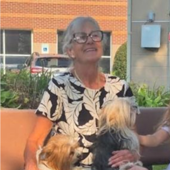 Woman with glasses smiles, holding two small dogs outdoors.