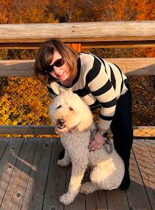 Woman in sunglasses petting a fluffy cream-colored dog on a wooden bridge with fall foliage in the background.