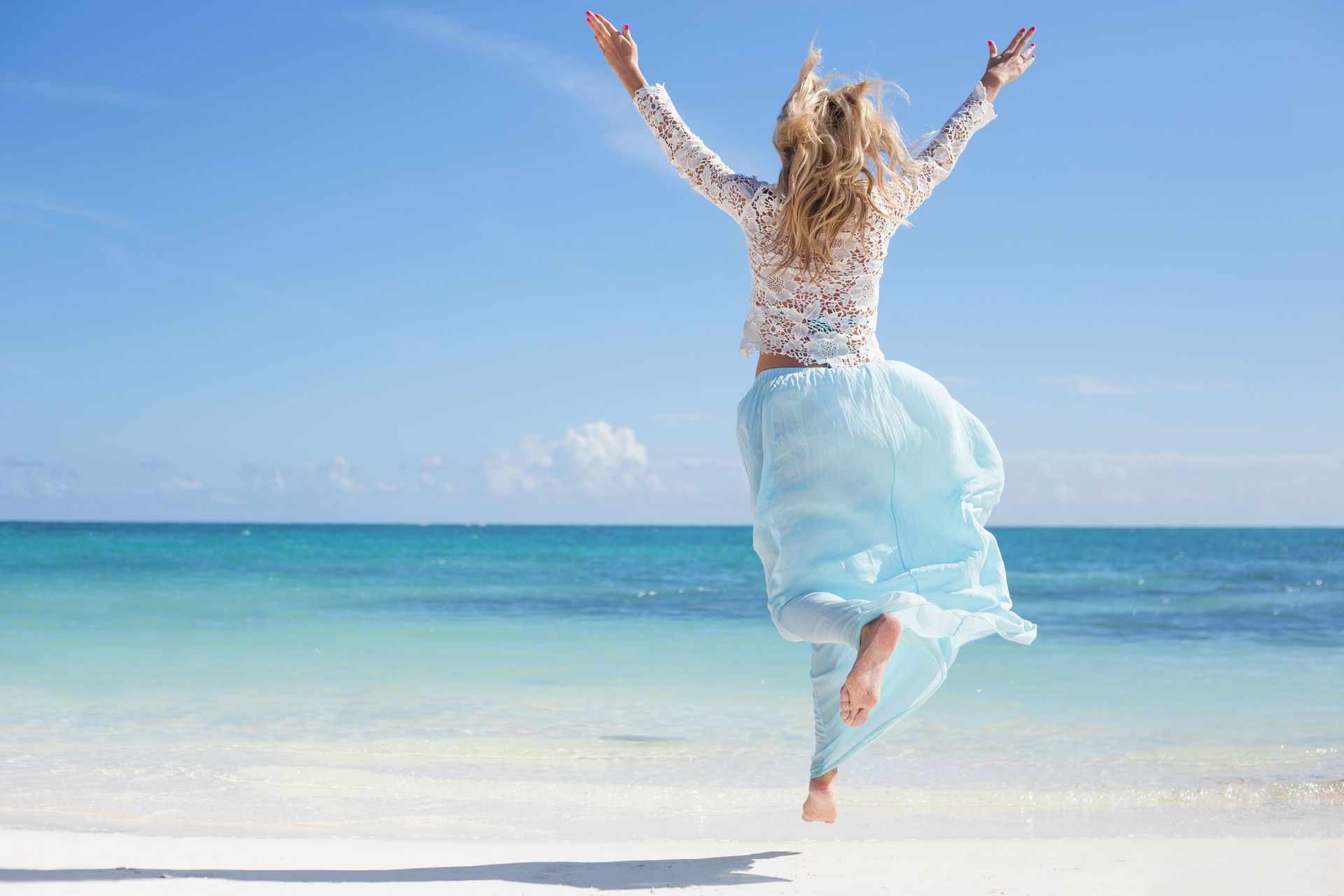 A woman in a blue skirt is jumping in the air on the beach.