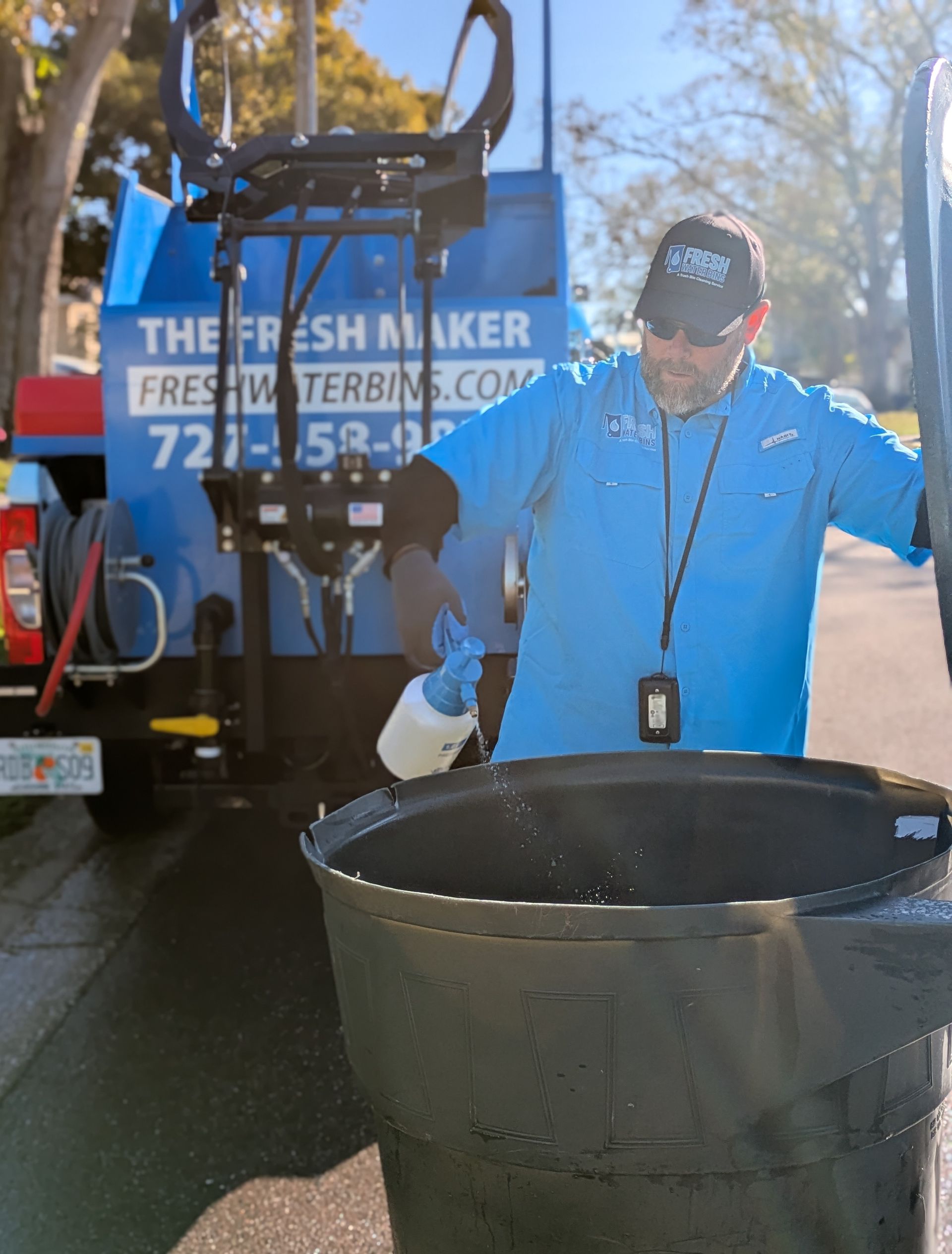 A man is spraying a green trash can with a spray bottle.