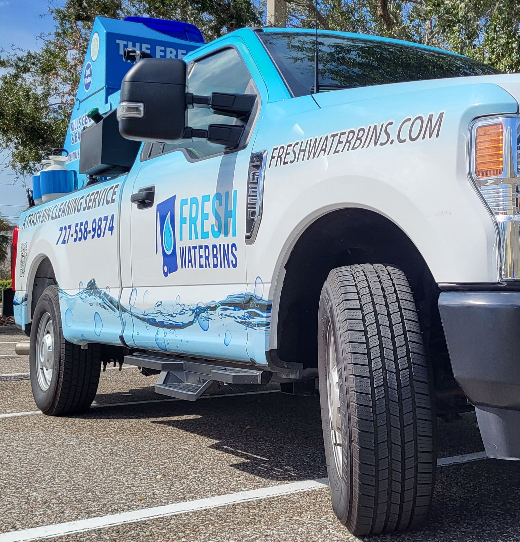 A green trash can with wheels is sitting on the sidewalk.
