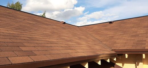 A close-up of a brown, shingled residential roof with a valley, partially visible rafters, and a blue sky above.