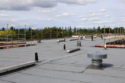 A flat commercial roof with grey membrane, ventilation pipes, and safety scaffolding against a cloudy blue sky.