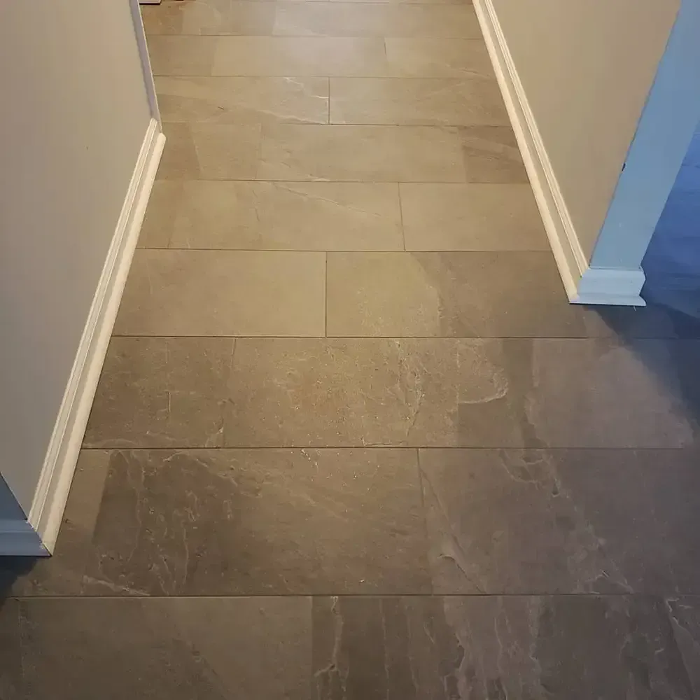 Gray tile floor in a hallway, bordered by white baseboards and gray walls.