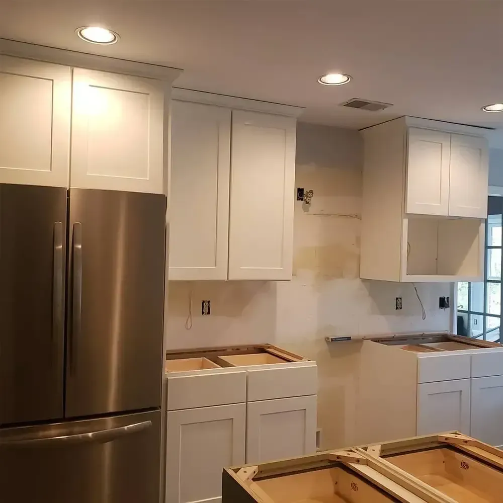 A kitchen under renovation with white cabinets, stainless steel refrigerator, and exposed drywall. The lower cabinets are installed, but countertops are missing.
