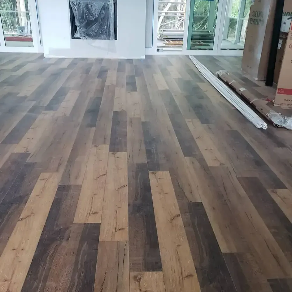 Newly installed wood-look flooring in a room with a fireplace and large windows. The floor has alternating light and dark brown planks.