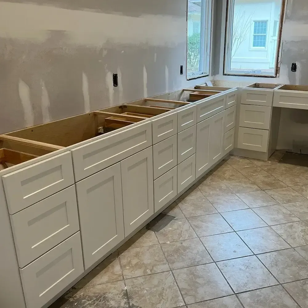 White kitchen cabinets installed against a wall, with open spaces above for countertops. Tile floor, two windows visible in the background.
