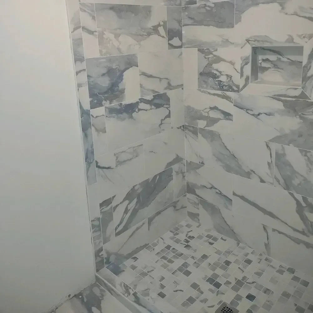 A shower interior with marble-patterned wall tiles in shades of gray and white, contrasting with the small mosaic floor tiles.