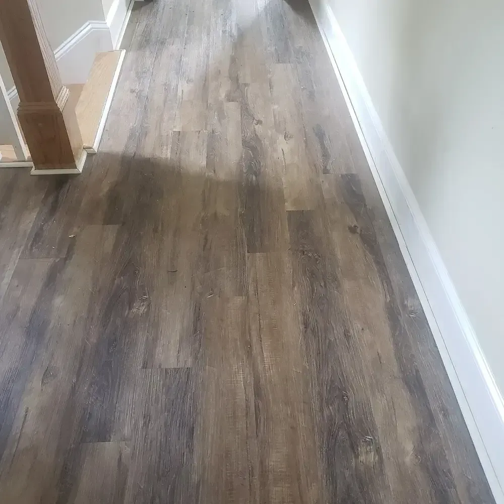 Hardwood-style flooring in a hallway, with white trim and a section of stairs visible. The floors are shades of brown and gray.