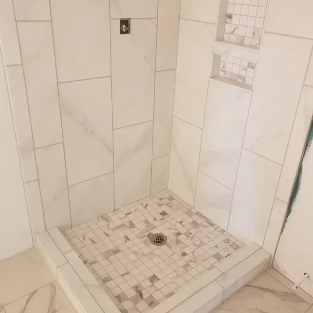 A newly tiled shower with white marble-look tiles and mosaic floor, with a built-in shelf.