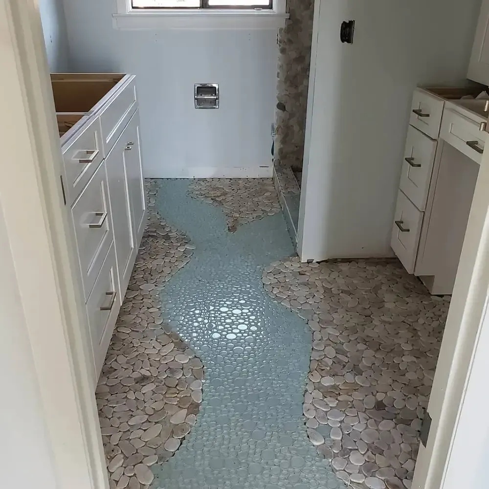 Bathroom with pebble tile flooring. A blue tile river flows through the beige pebble tile. White cabinets are on either side.