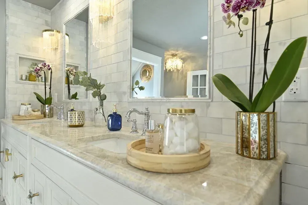 Elegant white bathroom with marble countertop, gold accents, and orchids. Features double sinks, mirrors, and decorative accessories.