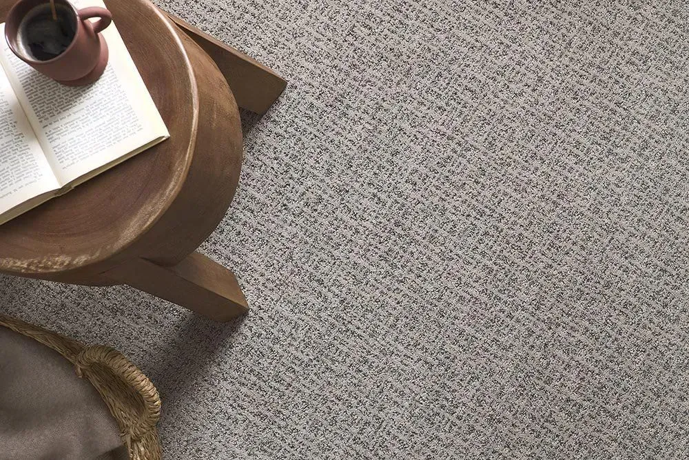 A round wooden side table with a book and mug, set on a patterned gray carpet.