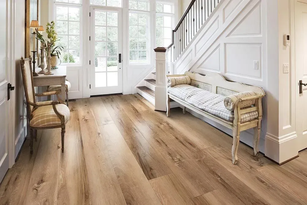 A bright entryway with light wood flooring, a white staircase, and a bench. A vintage chair sits near a table with decor.