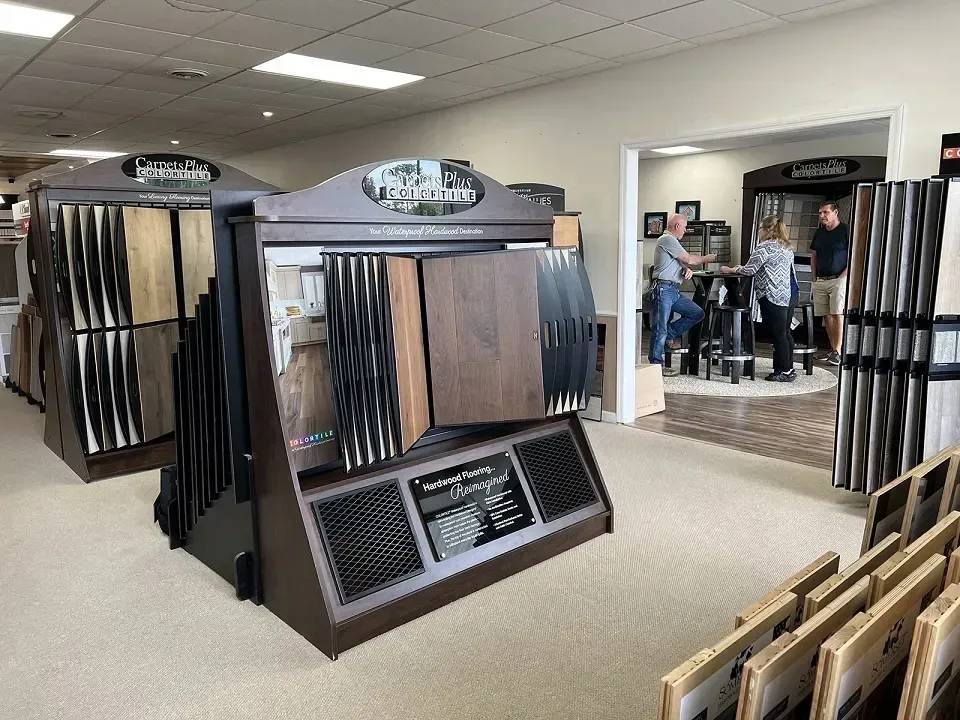 A flooring showroom with sample displays. Customers and a salesperson are visible in a back room, browsing flooring options. Beige carpet and wood samples are prominent.