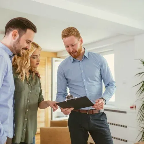 A real estate agent showing a young couple a document in a bright room. The agent points, and the couple looks on.