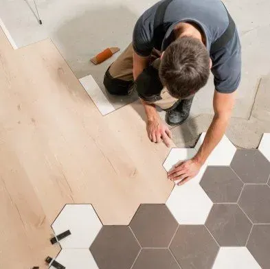 A worker installs hexagonal tiles on a floor, with both light wood and gray/white tiles.