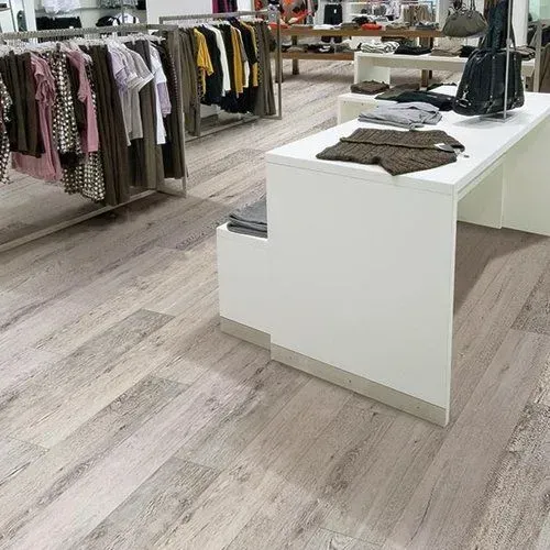 Inside a clothing store with light wooden floors. Clothes hang on racks. A white counter is in the center.
