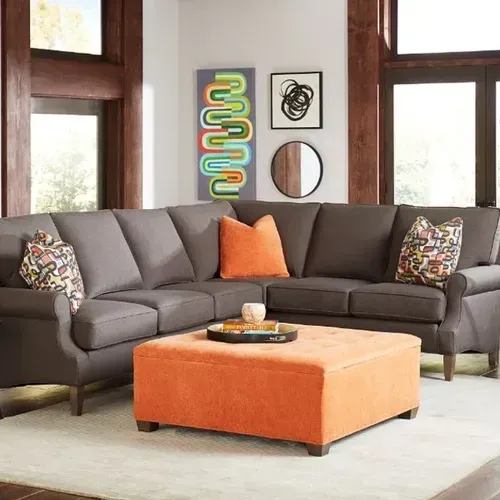 Gray sectional sofa in a living room, with an orange ottoman and throw pillows. Art and a mirror are mounted on the wall.