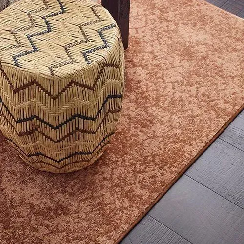 Woven stool with zigzag pattern next to a textured reddish-brown rug on dark wooden floor.