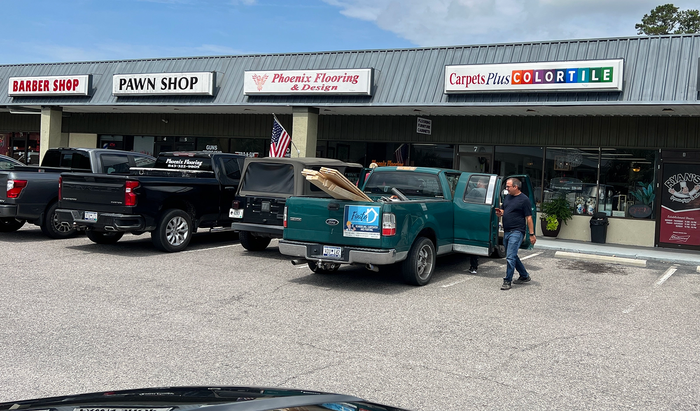 A man walks near a green pickup truck loaded with wood in front of a strip mall with various businesses.
