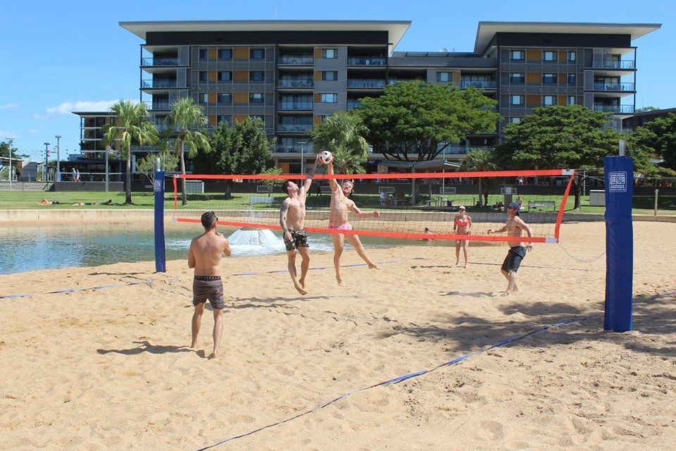 A group of people playing beach volleyball. A man in the back is jumping to spike the ball across the net
