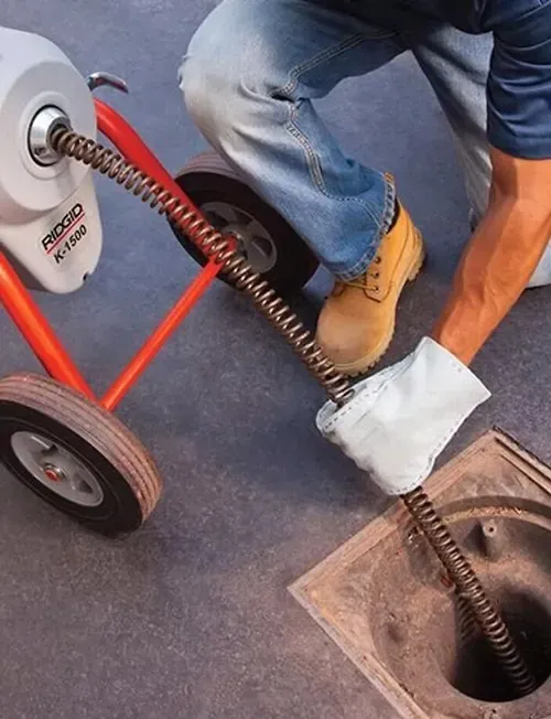 A person in work boots uses a RIDGID K-1500 drain cleaning machine to feed a cable into a ground-level sewer cleanout.