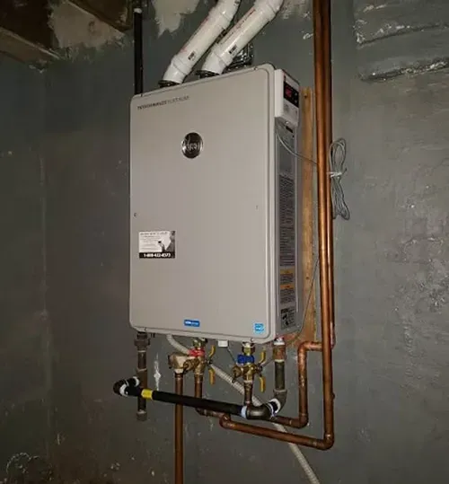 A grey, wall-mounted tankless water heater installed in a basement, connected to white exhaust pipes and copper plumbing.