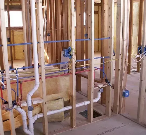 Interior wall framing under construction with exposed PVC plumbing pipes, electrical wiring, and insulation.