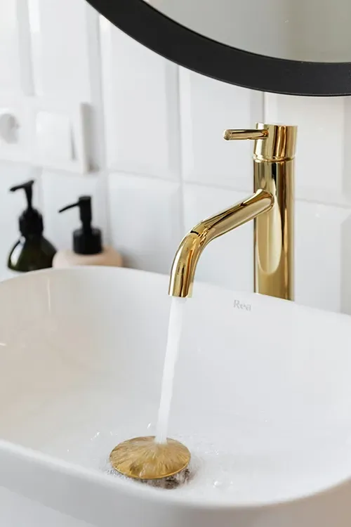 A gold-finished faucet pours water into a white vessel sink in a modern bathroom with white subway tile.