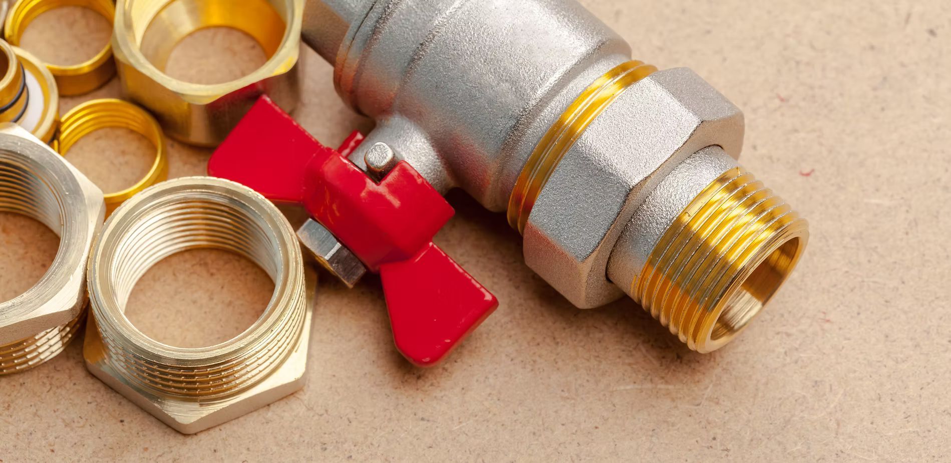 A metal ball valve with a red handle resting on a surface alongside several brass pipe fittings and connectors.