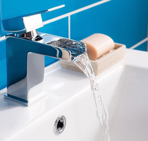 A modern chrome waterfall faucet pours water into a white sink, with a bar of soap on a tray nearby against blue tiles.