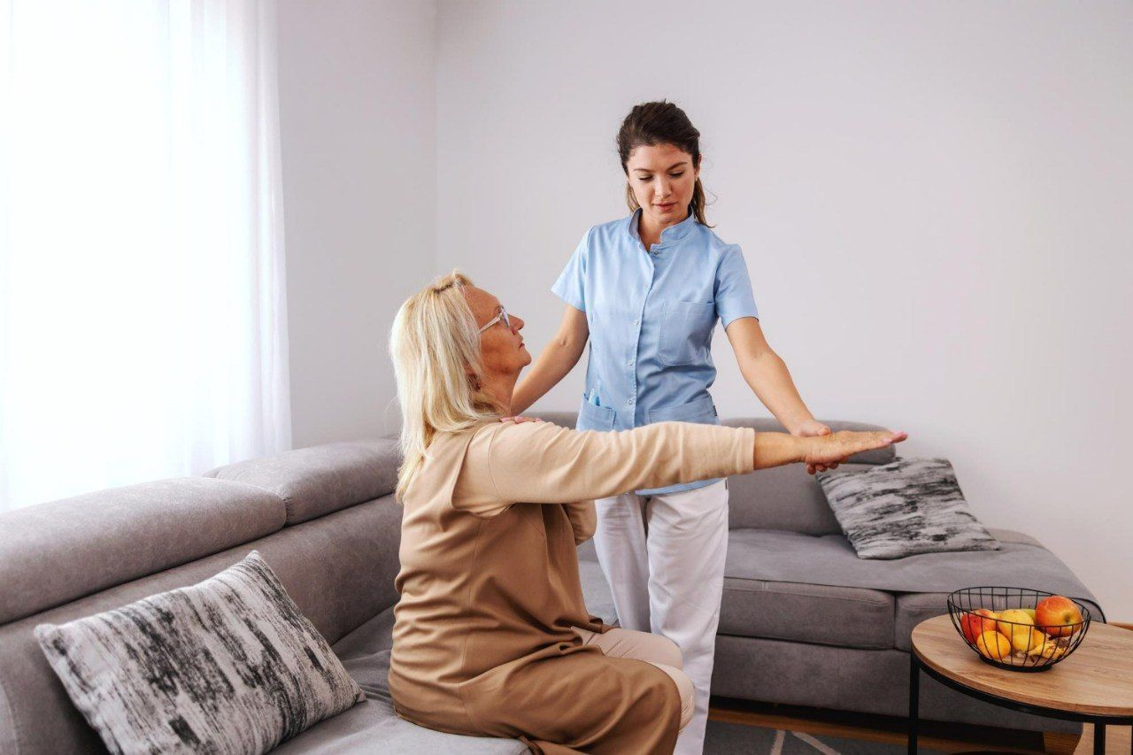 A healthcare worker assists a senior woman with arm exercises in a living room.