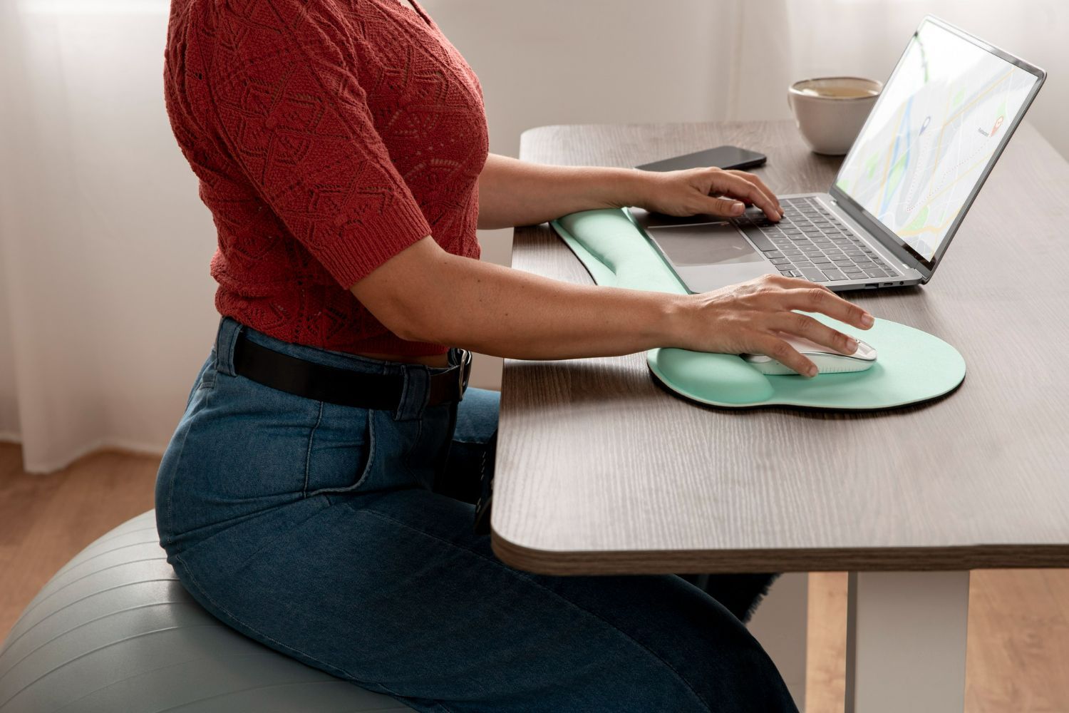 A woman is sitting on a yoga ball at a desk using a laptop computer.