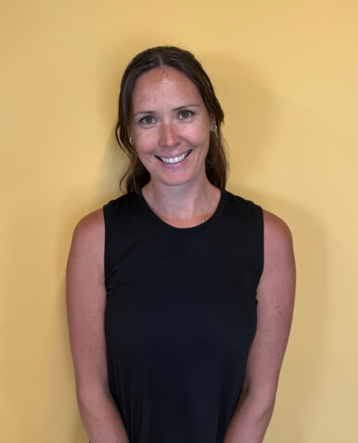 Woman smiling, wearing black tank top, standing in front of a yellow wall.