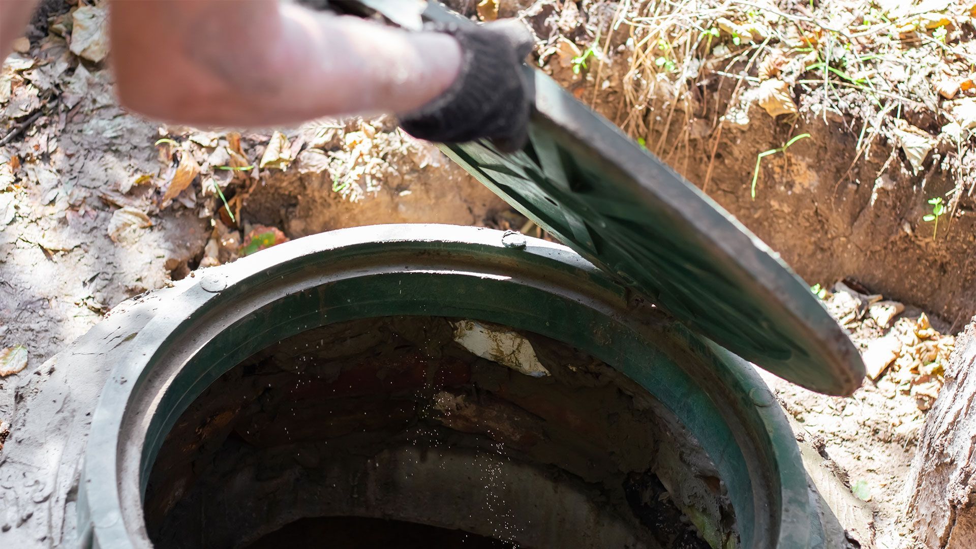 Person lifting the lid of a concrete septic tank, revealing wastewater inside. Outdoors, in a dirt setting.
