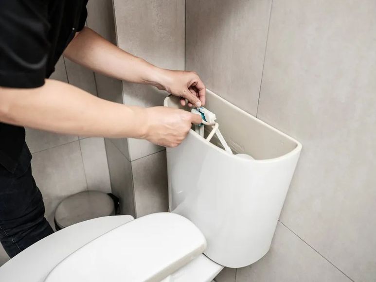 A person fixing a toilet's inner workings, holding a part within the white tank in a bathroom.