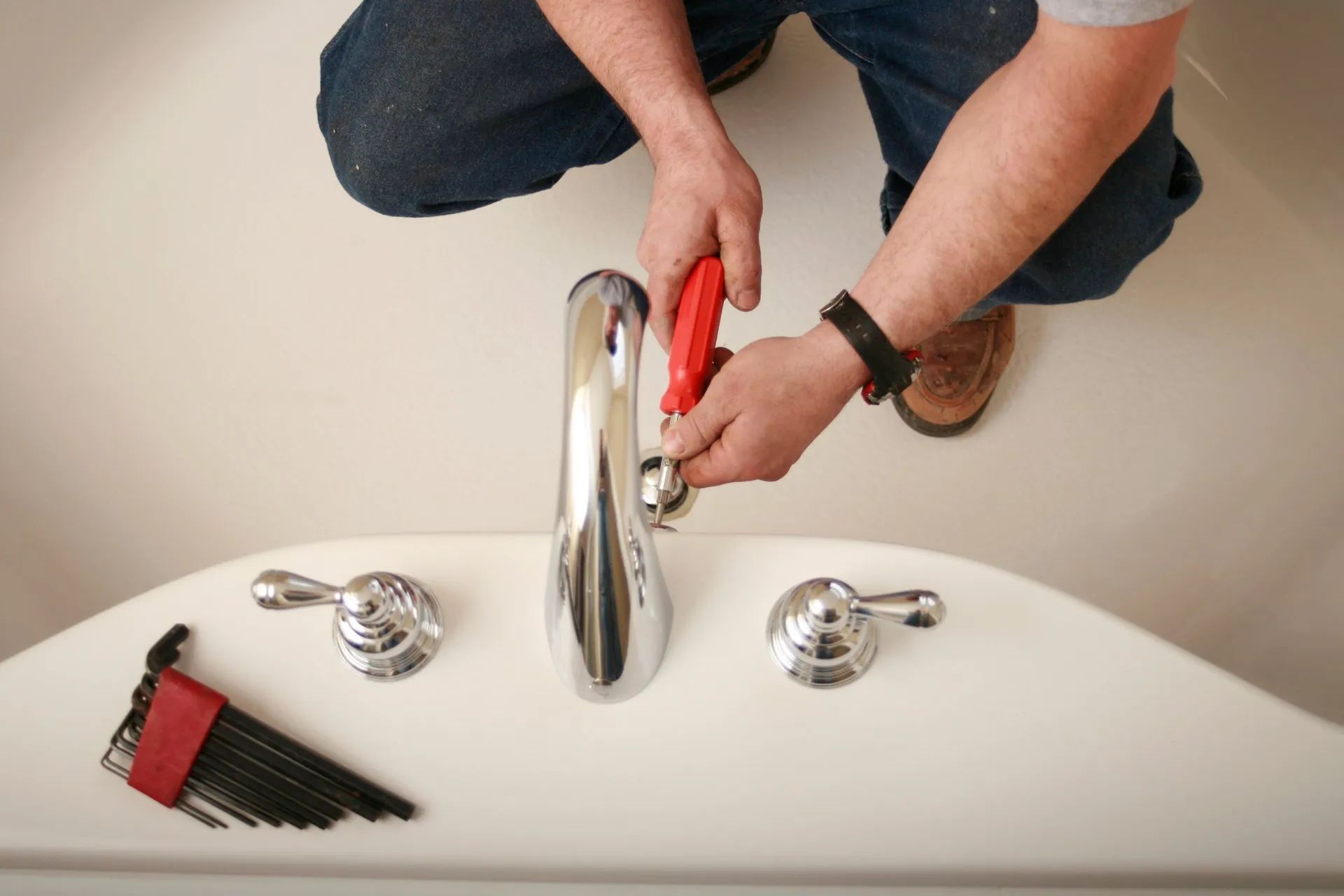 A person installing a faucet in a white bathtub, using a wrench. There are two handles and a set of Allen wrenches visible.