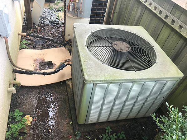 A dirty air conditioner is sitting on the ground next to a fence.