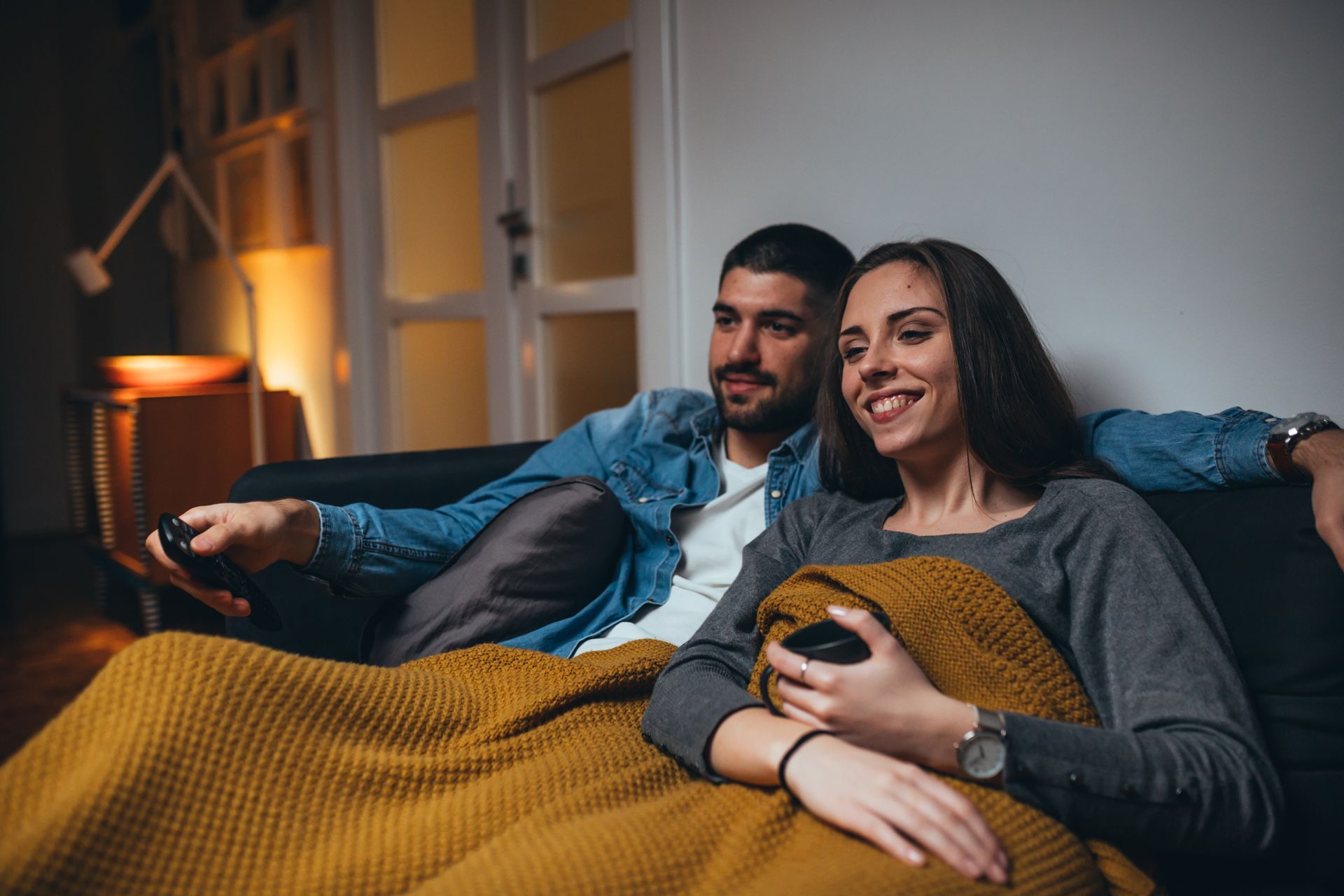Couple sitting on a couch, watching TV with remote. They are covered by a yellow blanket, smiling in a dimly lit room.