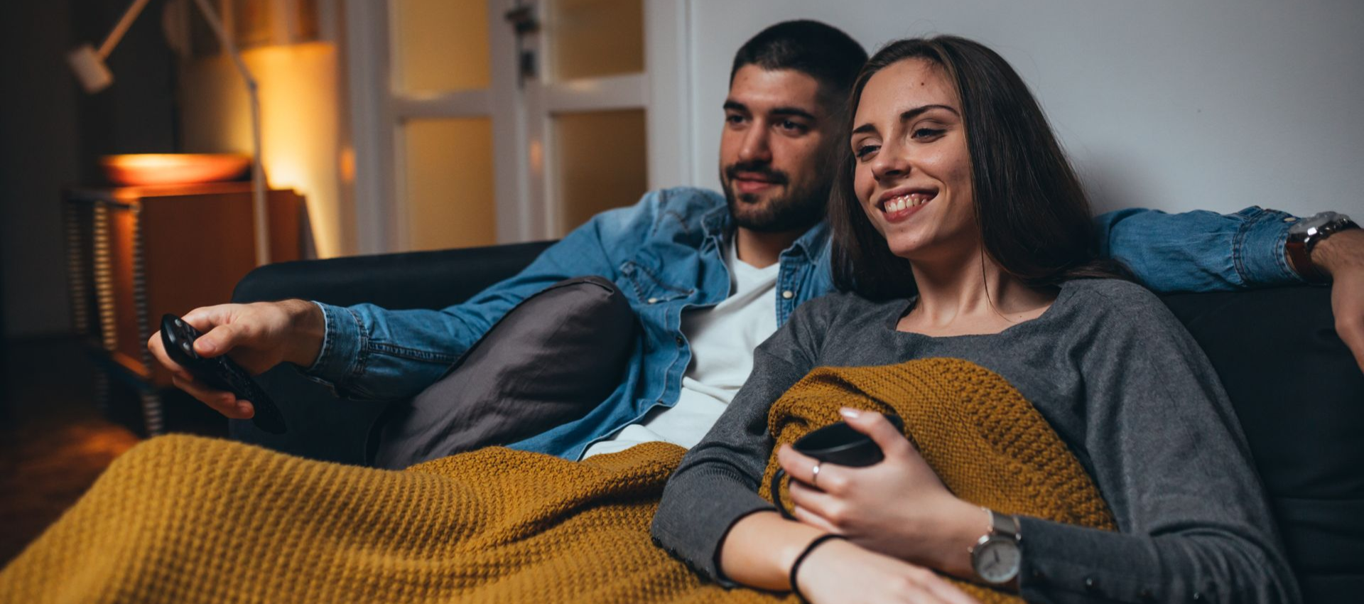 Couple on a couch watching TV, wrapped in a blanket, smiling. Dimly lit room.