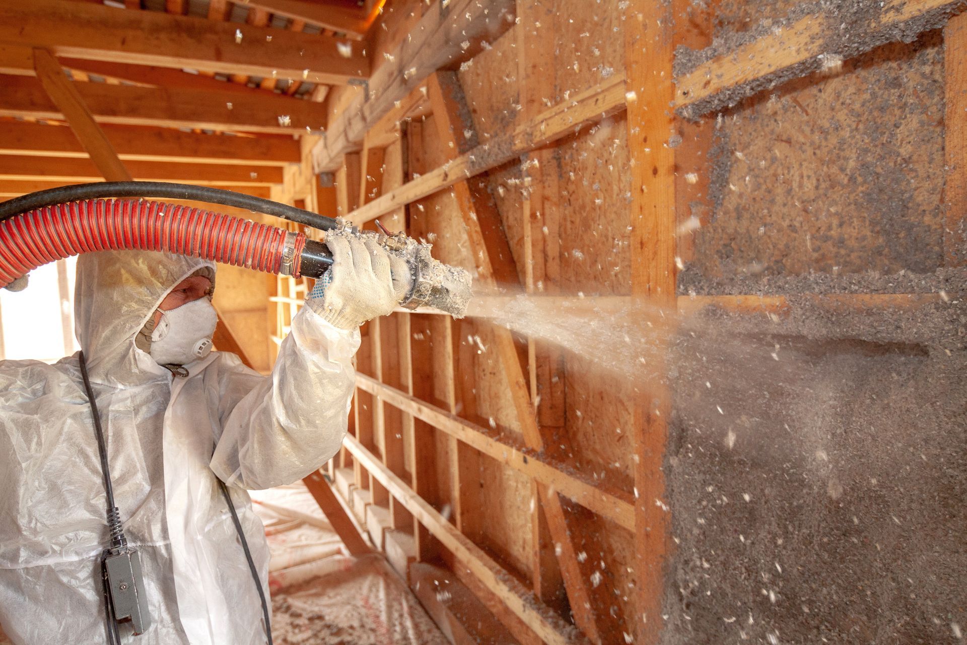 A man in a protective suit is spraying insulation on a wooden wall.