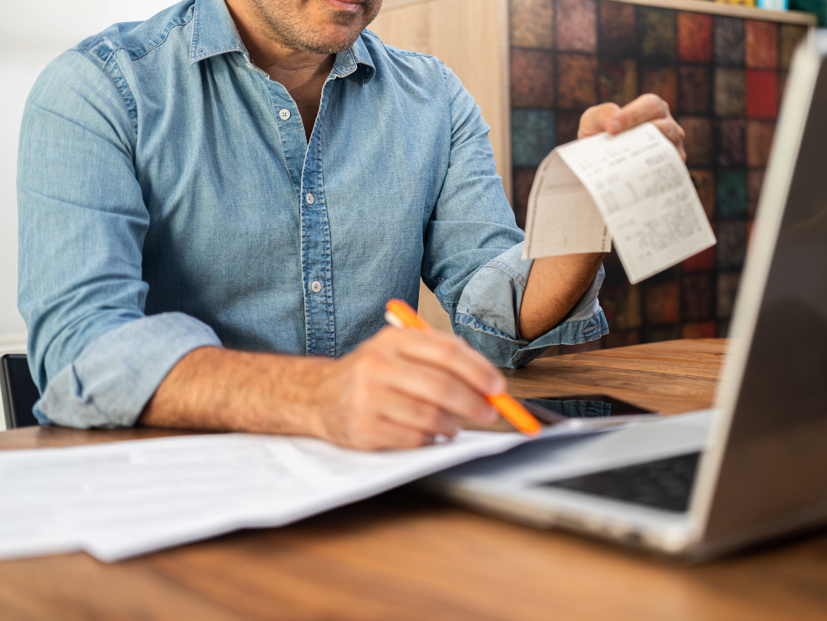Person writing on paper beside a laptop, holding a receipt at a desk.