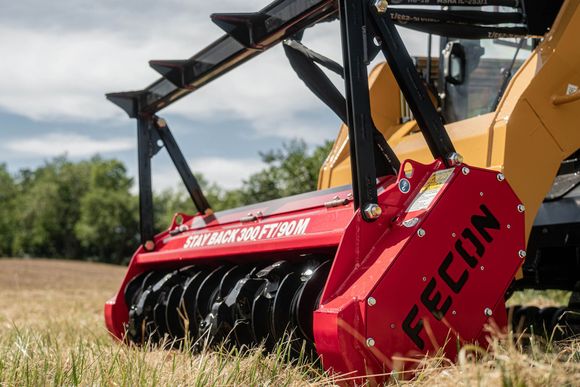 A red Fecon mulching head attachment on a yellow construction vehicle in a field, with a safety warning sign.