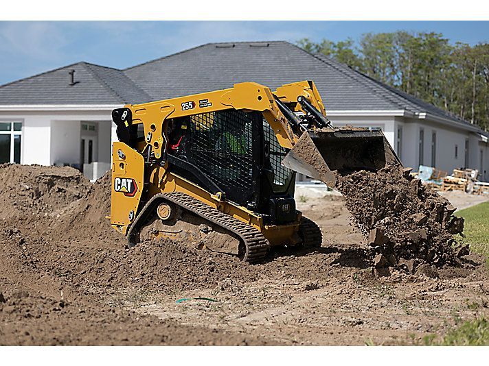 A yellow Caterpillar compact track loader moving a bucket of dirt at a construction site in front of a new house.