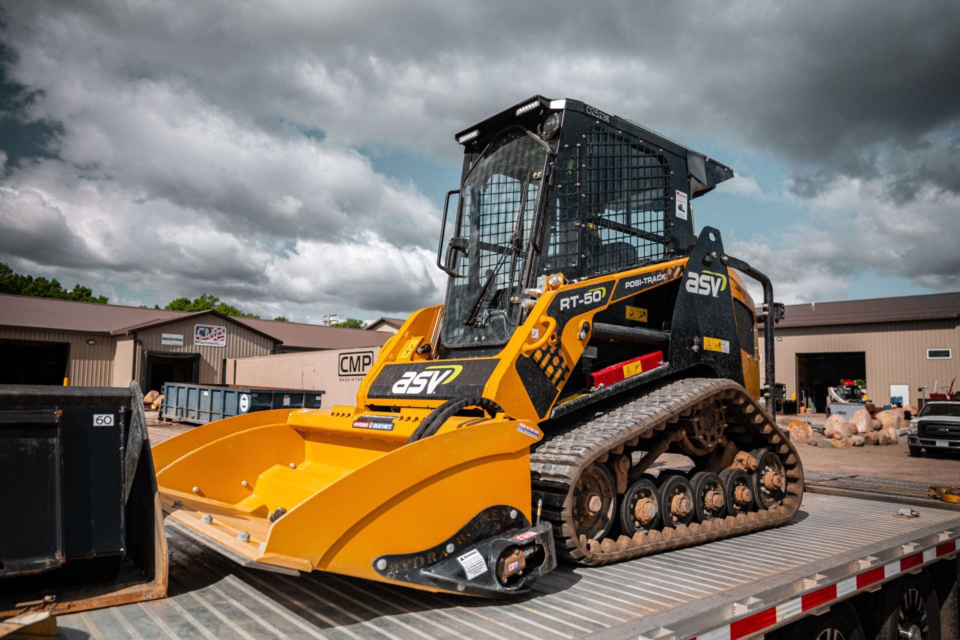 A yellow and black ASV compact track loader sits on a flatbed trailer in front of industrial buildings under a cloudy sky.