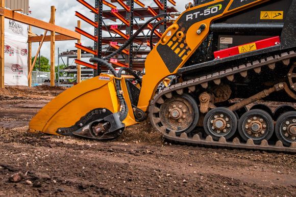 A yellow skid steer with tracks uses a stump grinder attachment to clear dirt at a construction site.