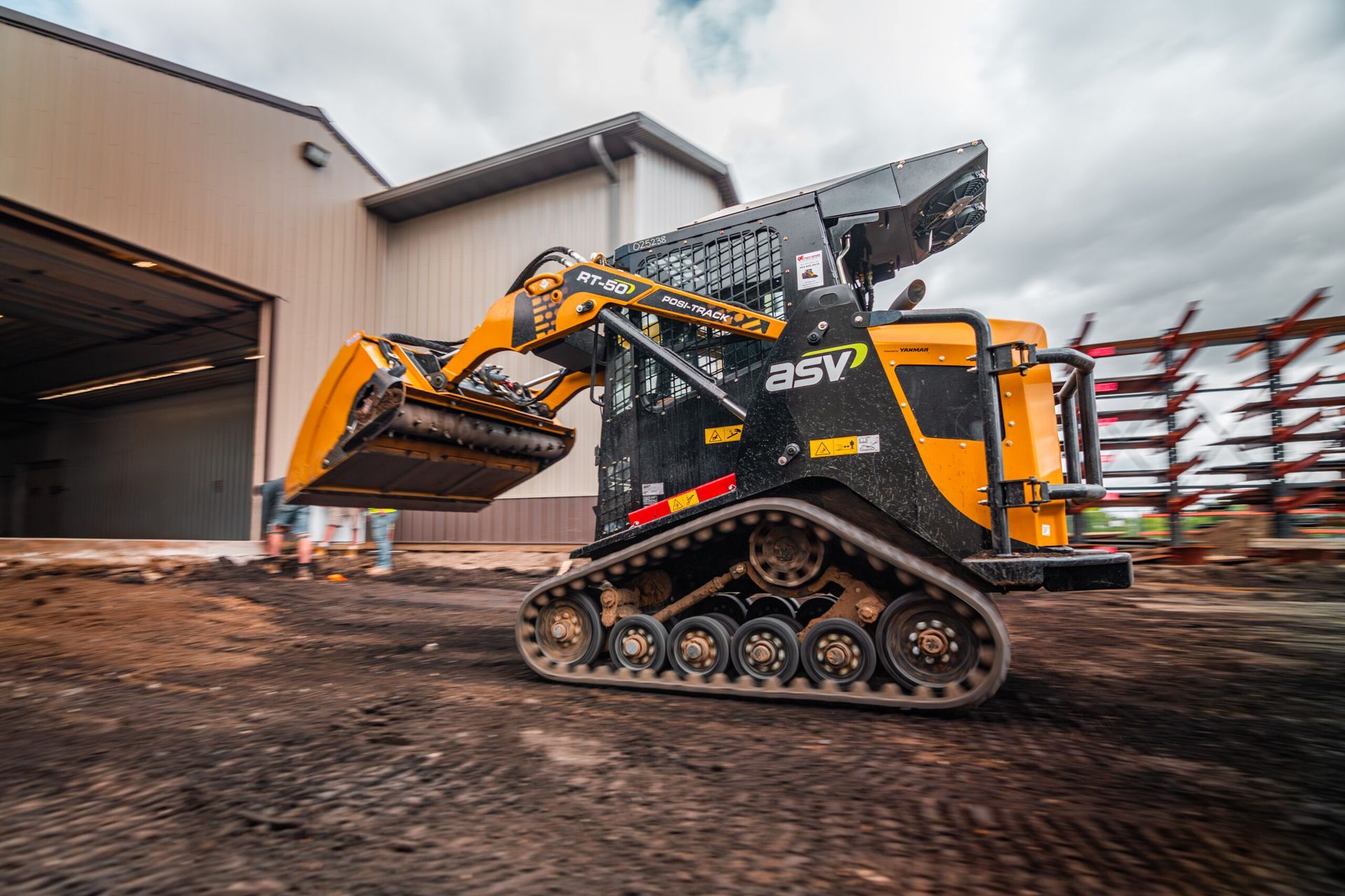 A yellow and black ASV compact track loader with a bucket attachment parked on dirt outside an industrial building.