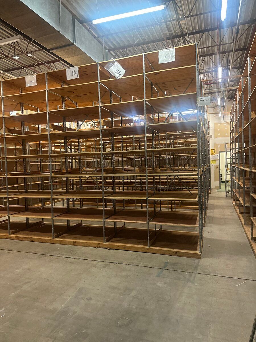Rows of empty, multi-tiered industrial wooden shelving units in a warehouse with high ceilings and concrete floors.